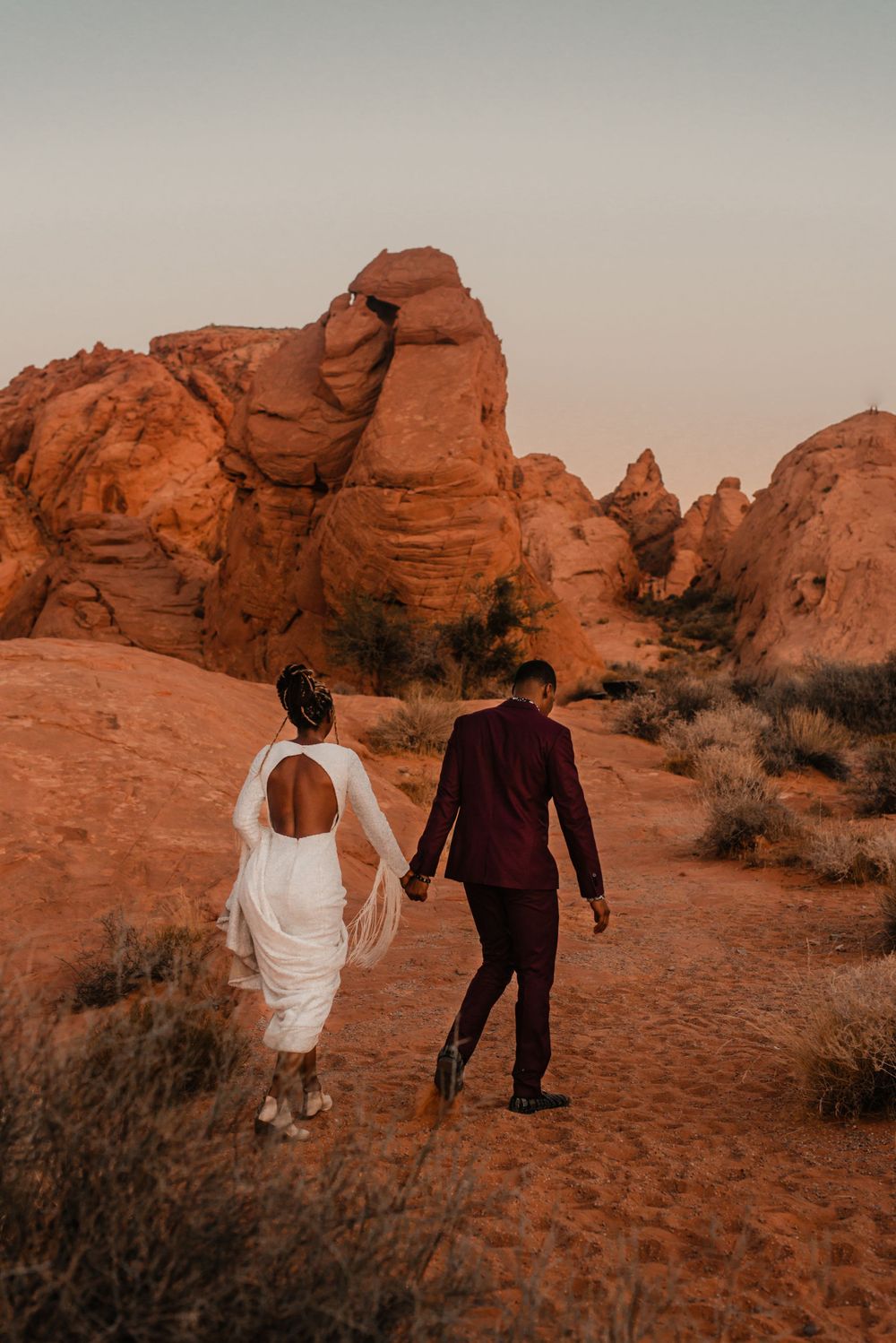 Couple walks hand in hand through red rock desert landscape wearing formal attire.