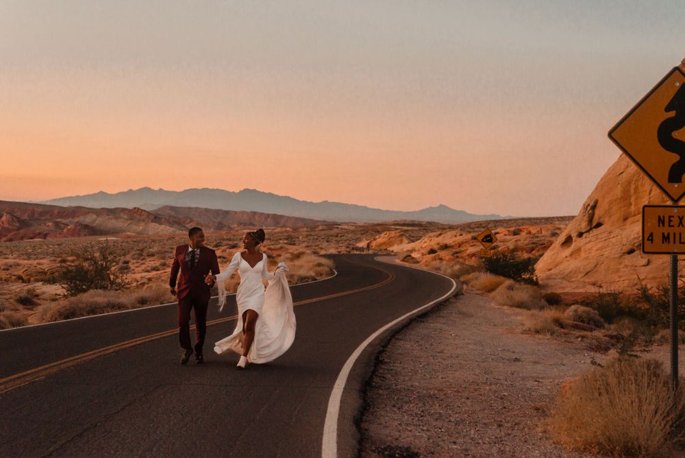A couple walking down a curved desert road at sunset with mountains in the distance.