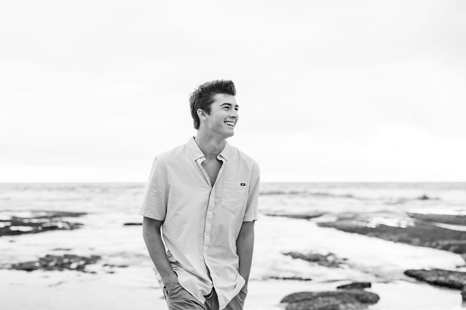 A high school senior in casual white shirt laughing while standing on rocky beach during photoshoot.