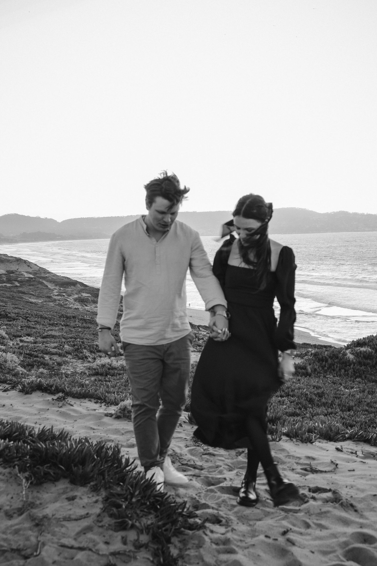 A couple walks hand in hand on a beach during a moody black and white photoshoot.