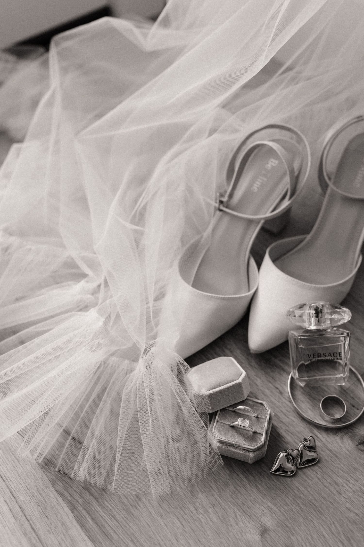 Wedding accessories including white heels, veil, rings and perfume bottle arranged on wooden surface.