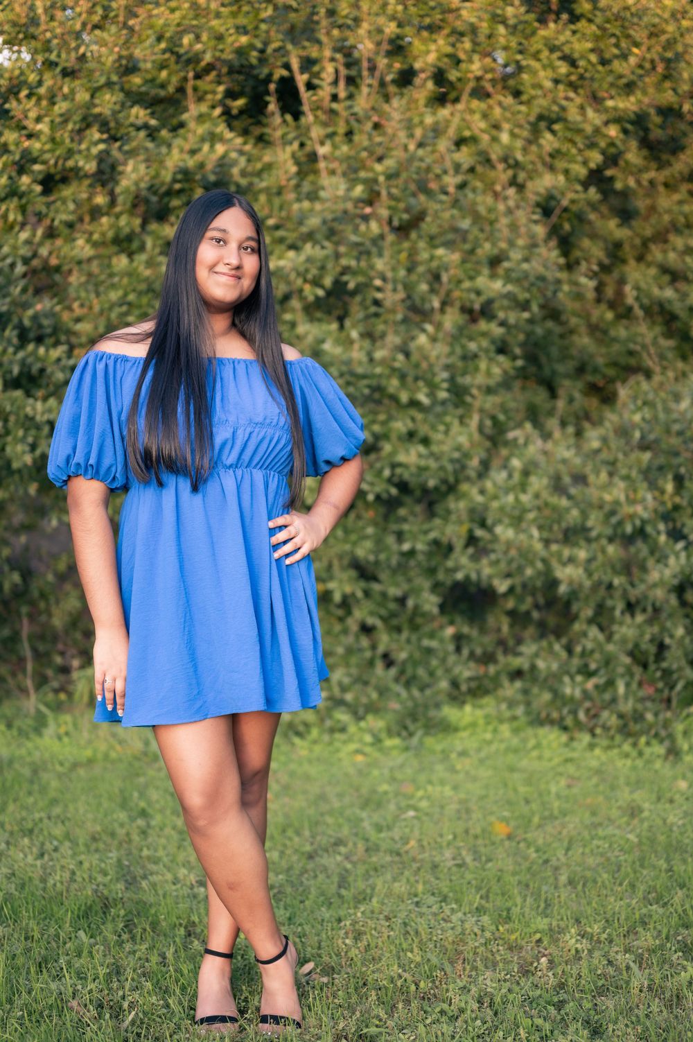 Woman in bright blue off-shoulder dress poses on grass with trees in background.