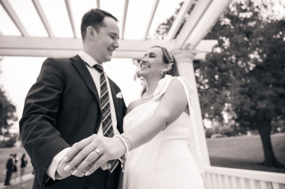 A black and white photo of newlyweds sharing a joyful moment under a pergola.