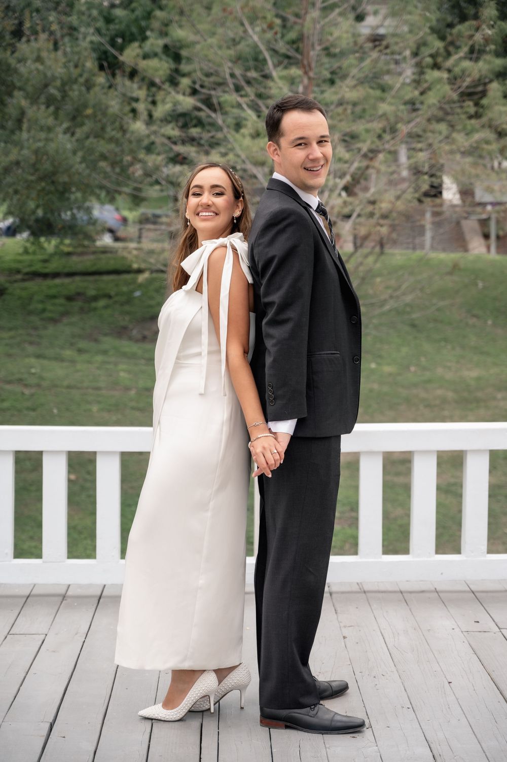 Bride and groom pose back-to-back on a white deck with scenic forest backdrop.