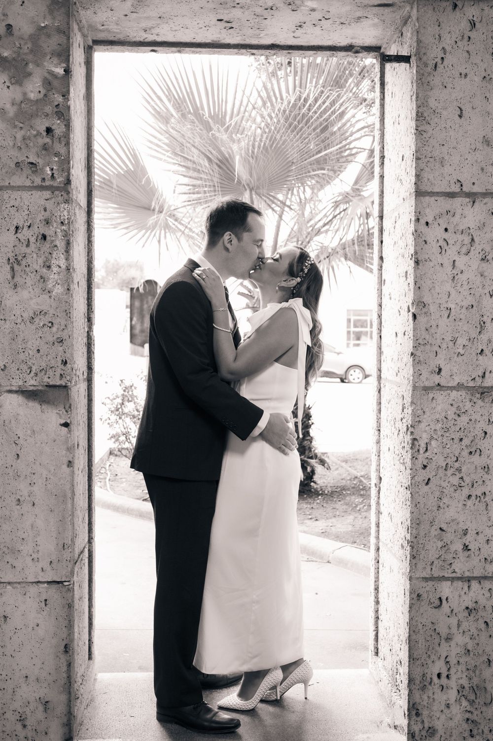 Black and white photo of couple embracing in stone doorway with palm trees visible.