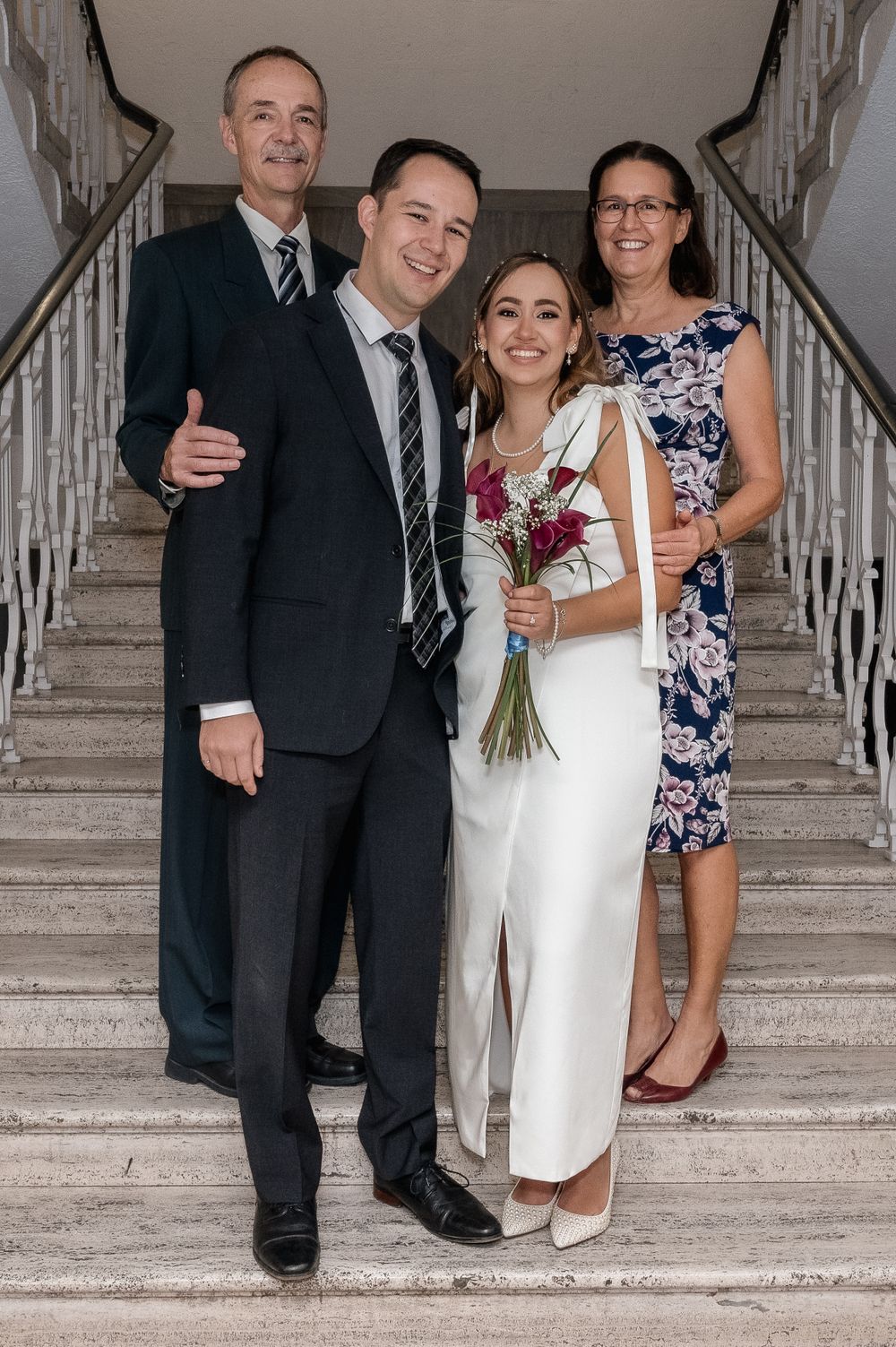 Wedding party poses on grand staircase with bride in white dress holding a burgundy bouquet.