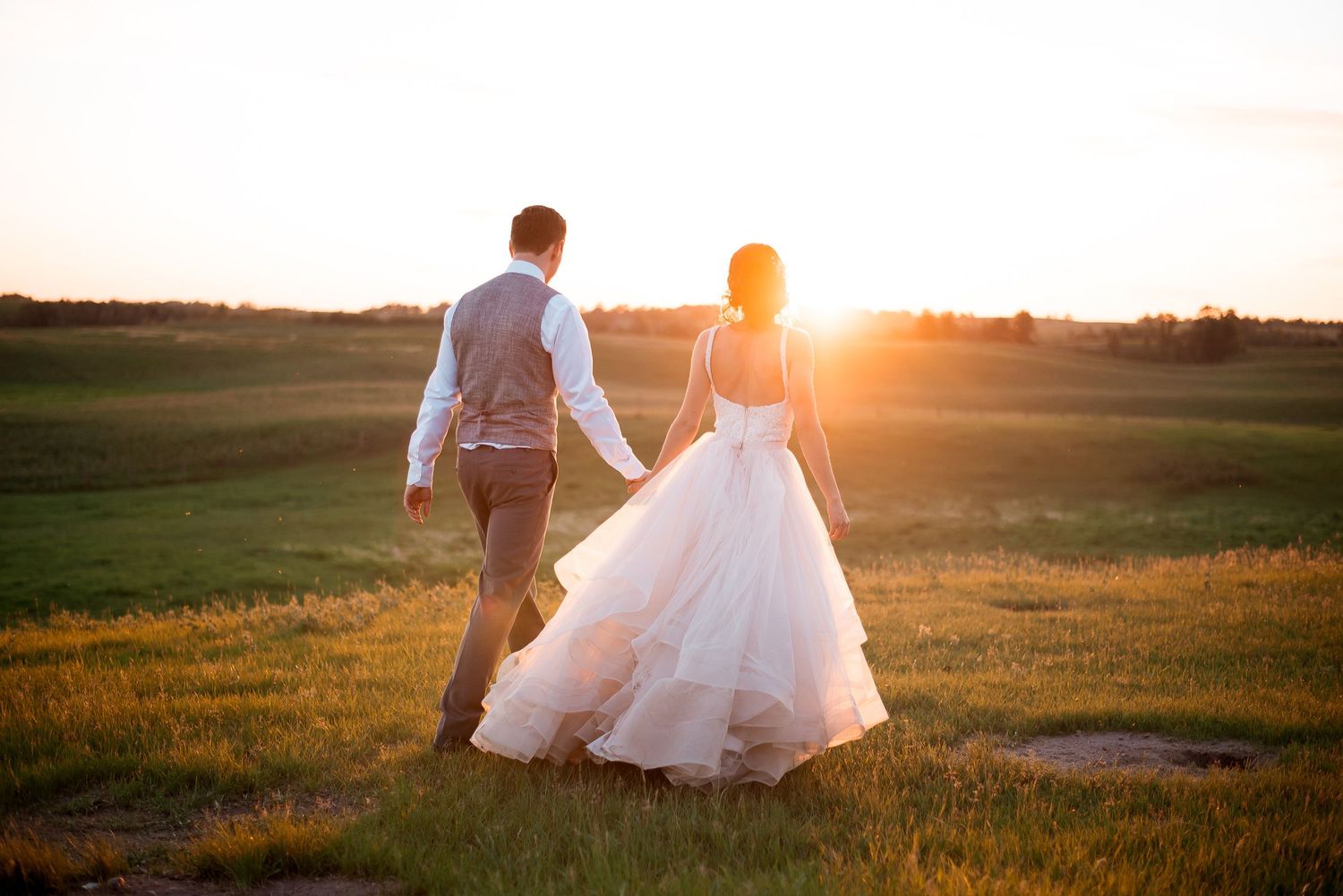 Newlywed couple walking through golden field at sunset with flowing wedding dress.