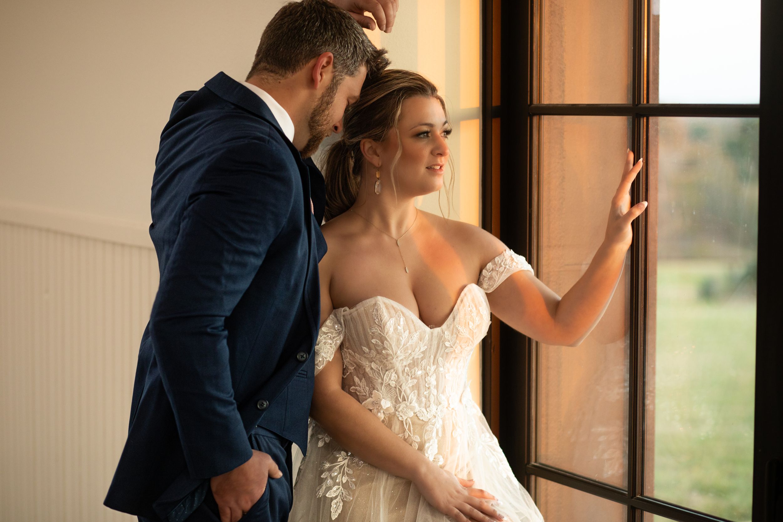 A couple in wedding attire stands intimately by a window in soft natural light.