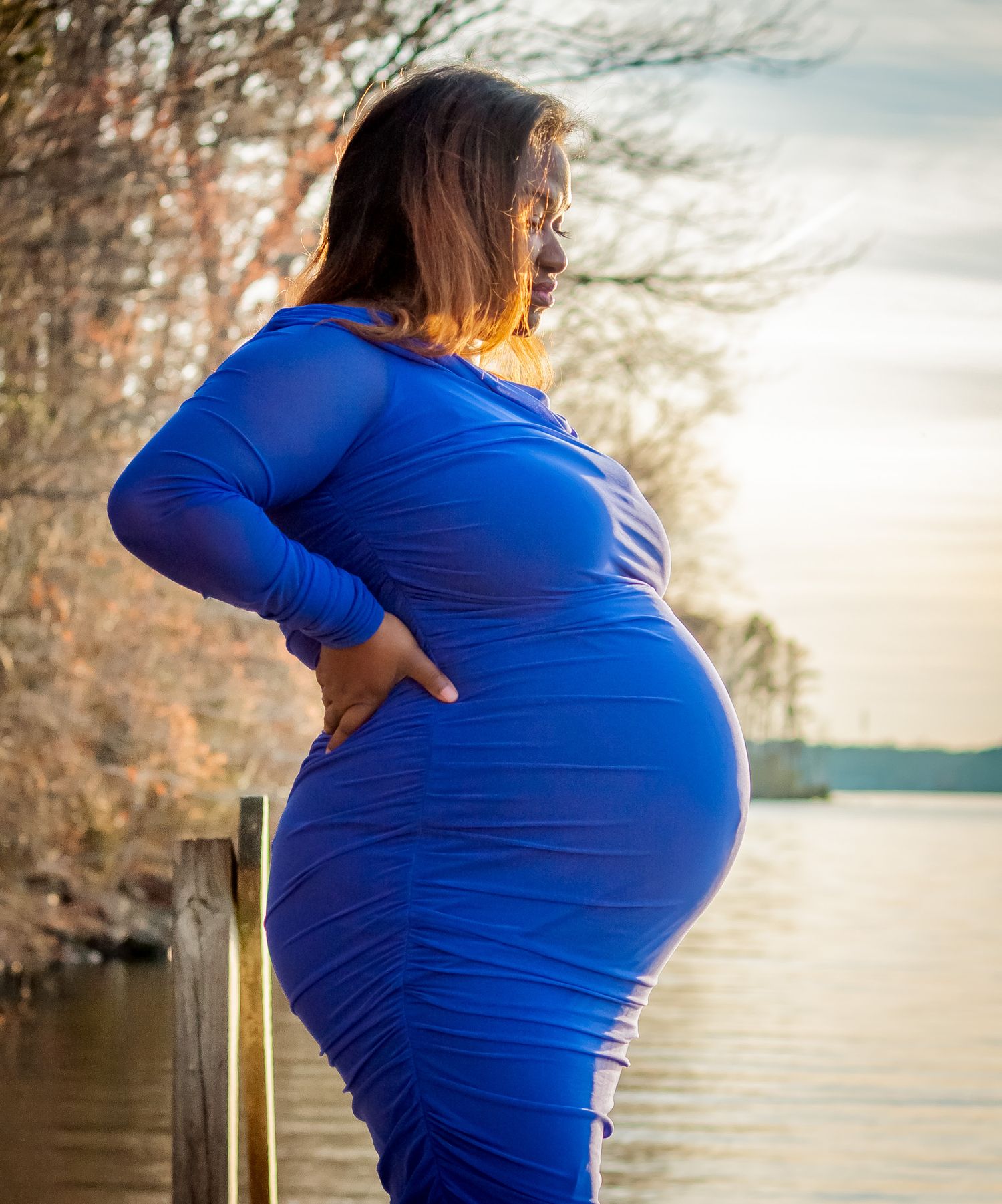 Silhouette of expectant mother in blue dress standing by waterfront at sunset.