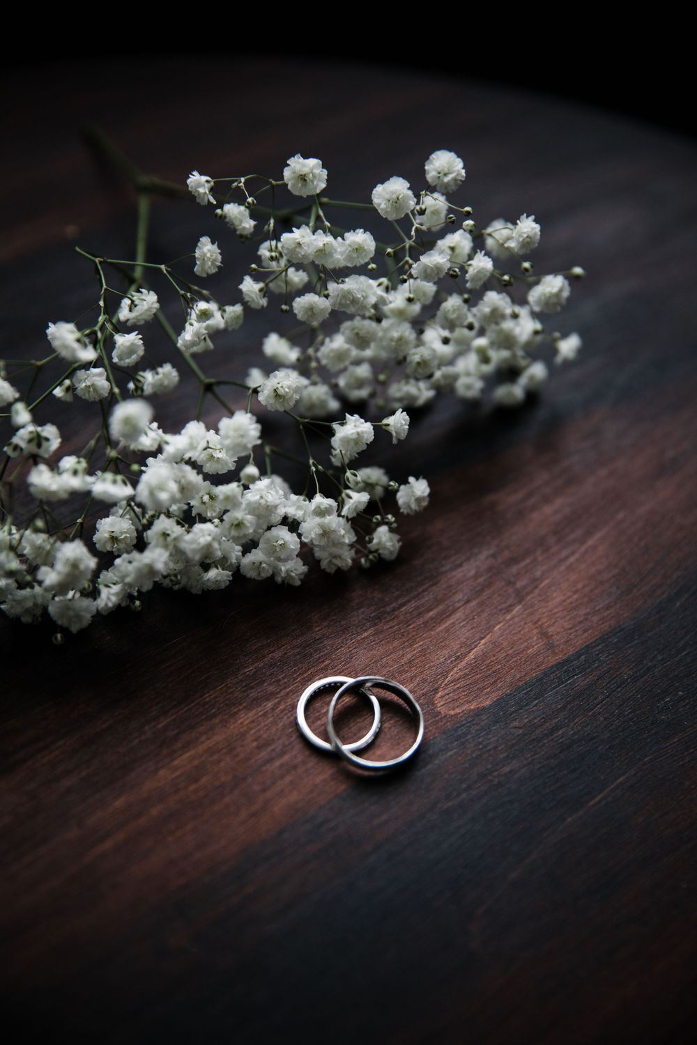 Wedding rings alongside delicate baby's breath flowers on dark wooden surface.