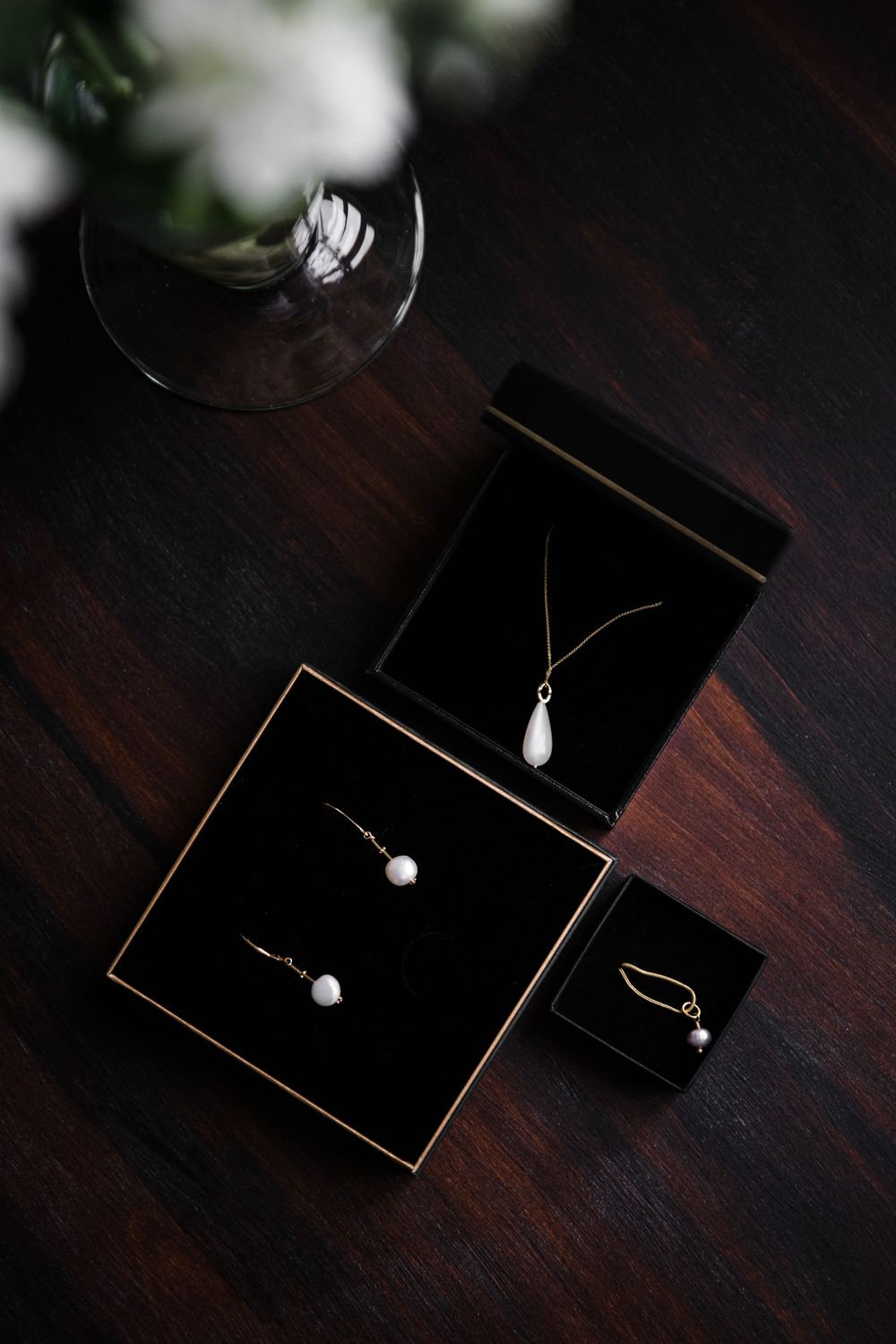 Pearl jewelry displayed in black boxes on dark wooden surface with wine glass.