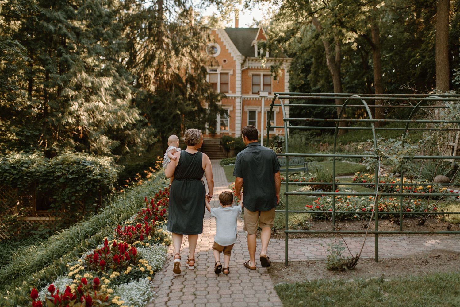 Family walking along garden path towards brick house with blooming red flowers.