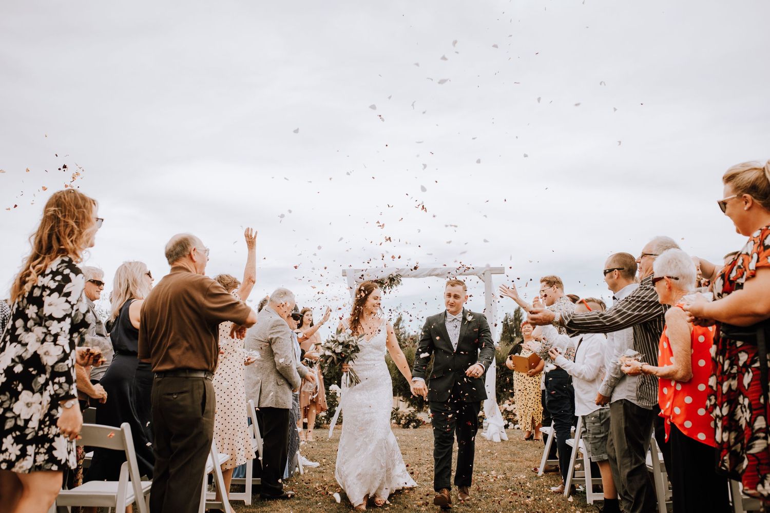 Wedding celebration scene with guests throwing confetti against cloudy sky.
