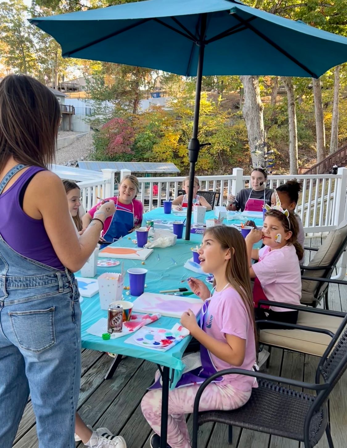 An outdoor paint party for a birthday with people painting at tables under blue umbrellas by a pool.