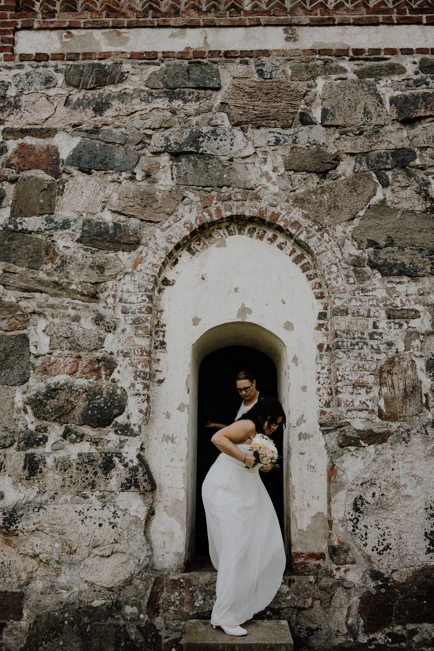 A figure in a white wedding dress poses in a stone archway of a historic building.