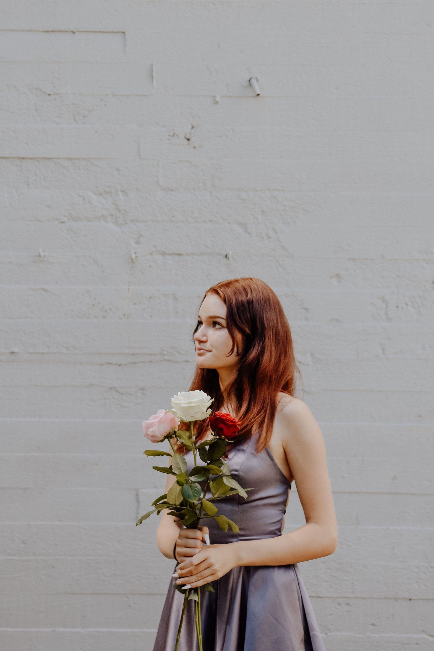 A person in a gray dress holding pink roses against a white brick wall.