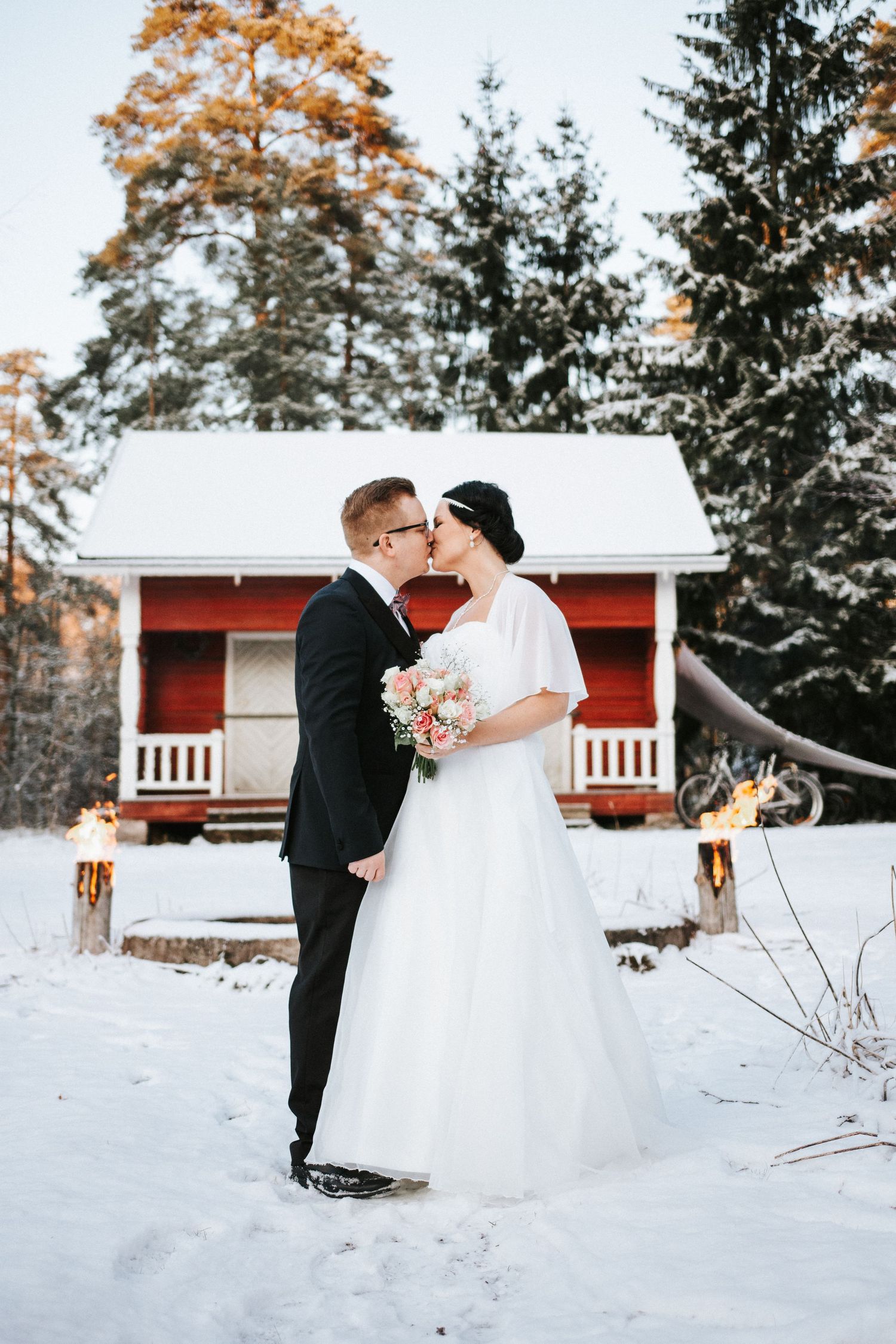 Couple sharing a romantic moment in front of a red cabin surrounded by snow and evergreen trees.