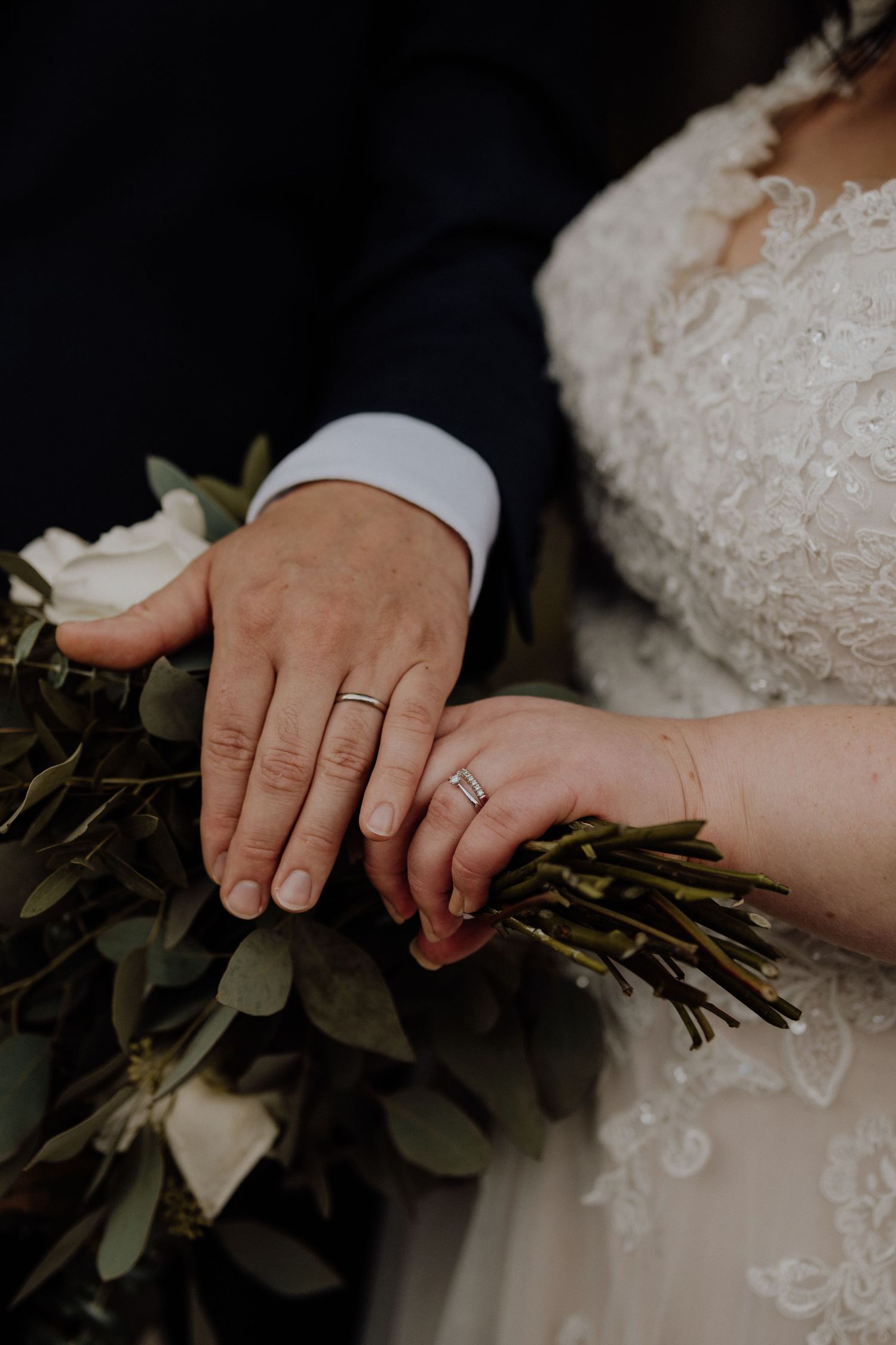 Close up of newlywed couple's hands with rings resting on a eucalyptus bouquet.
