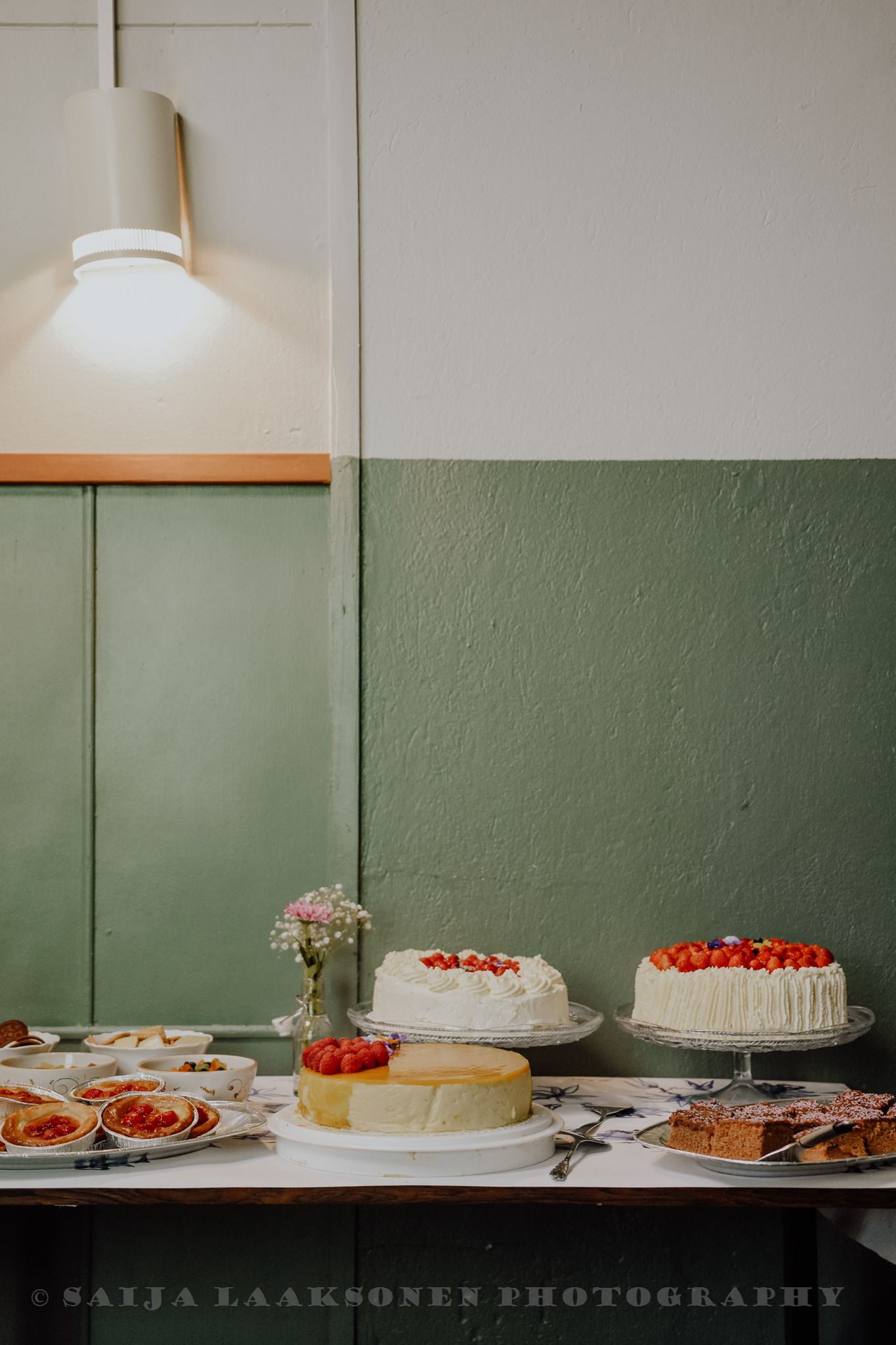 A dessert table featuring several elegant white cakes and pastries against a sage green wall.