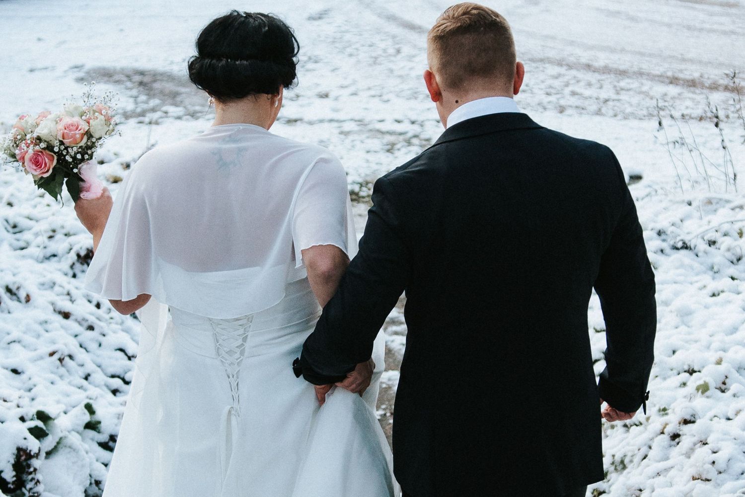 Back view of couple in wedding attire walking through snowy landscape.