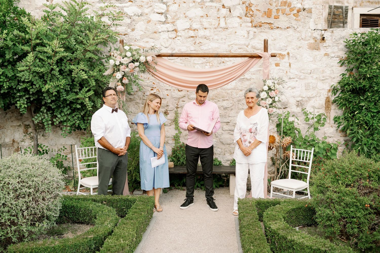 Wedding party members stand under pink draped arch in garden setting.