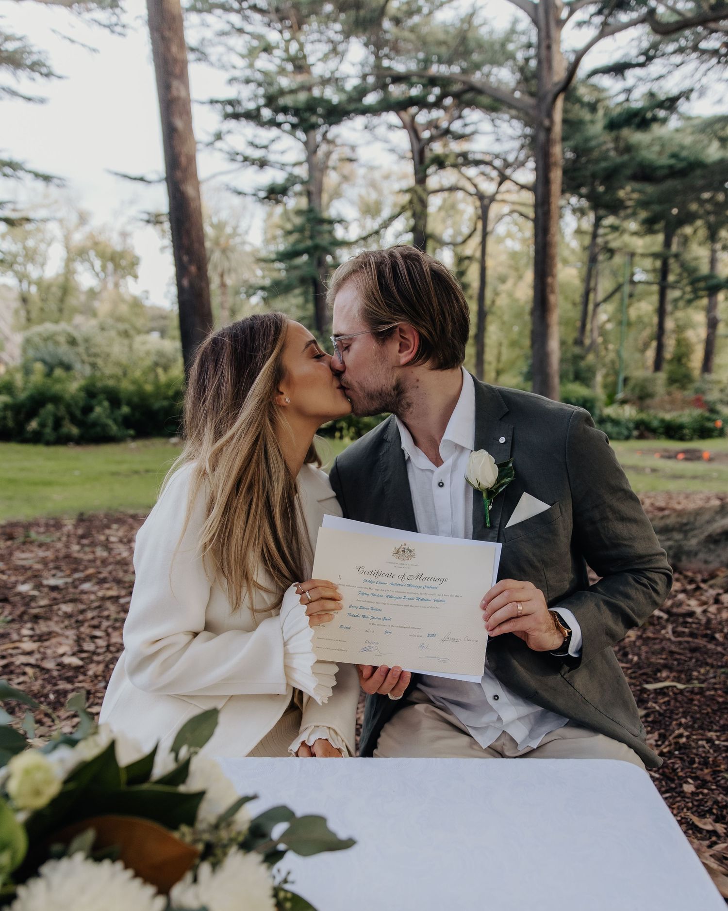 Newlyweds sharing a kiss while holding marriage certificates in an outdoor park setting.