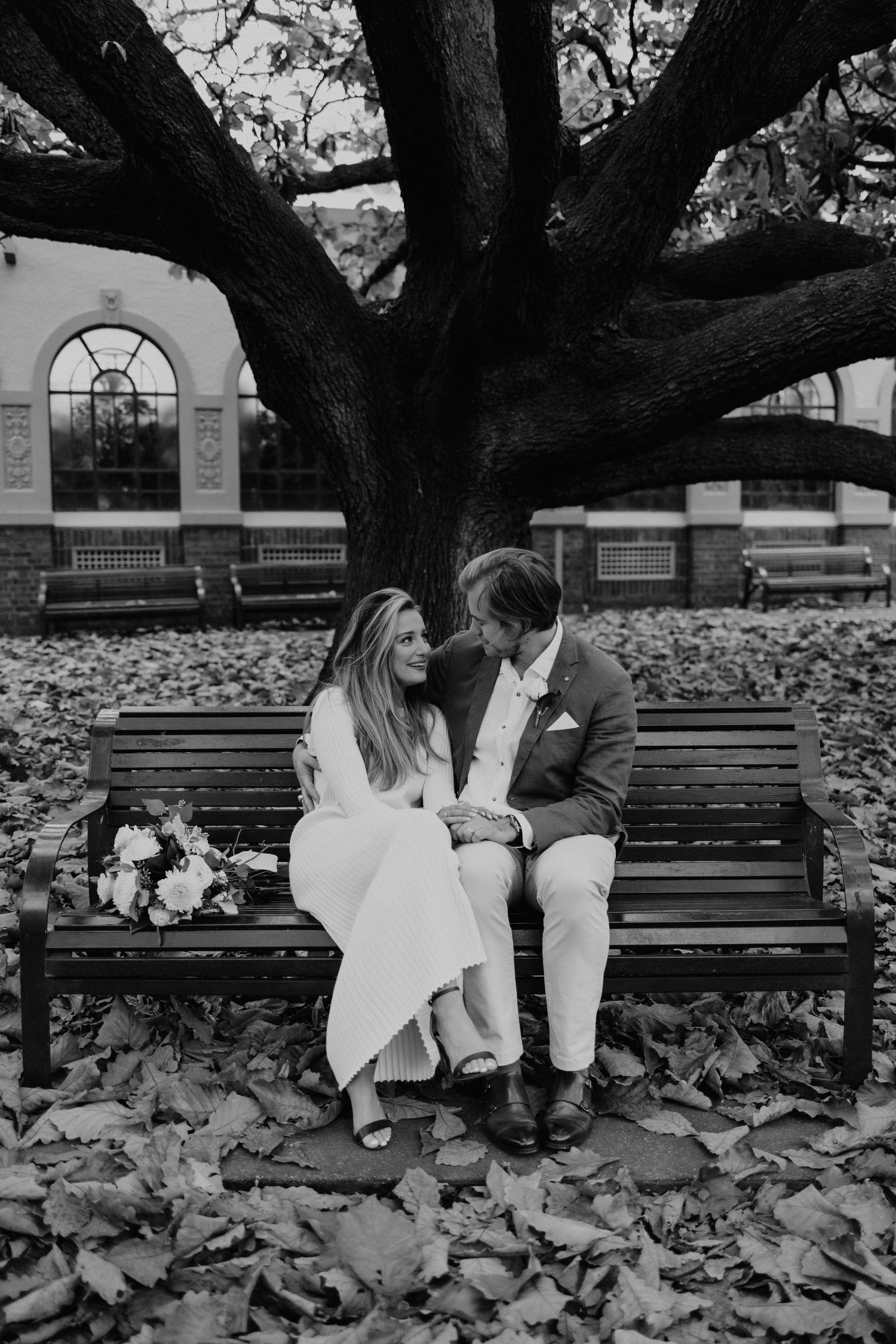 Couple seated on wooden bench surrounded by fallen autumn leaves in a park.