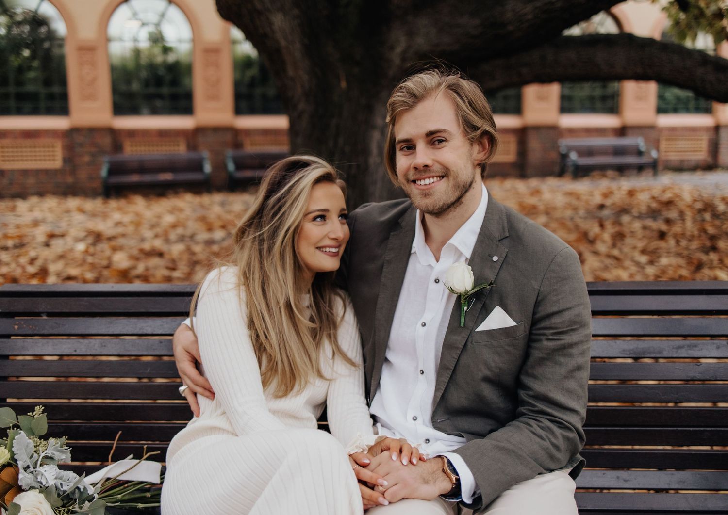 Newlyweds sharing tender moments on park bench near brick building.