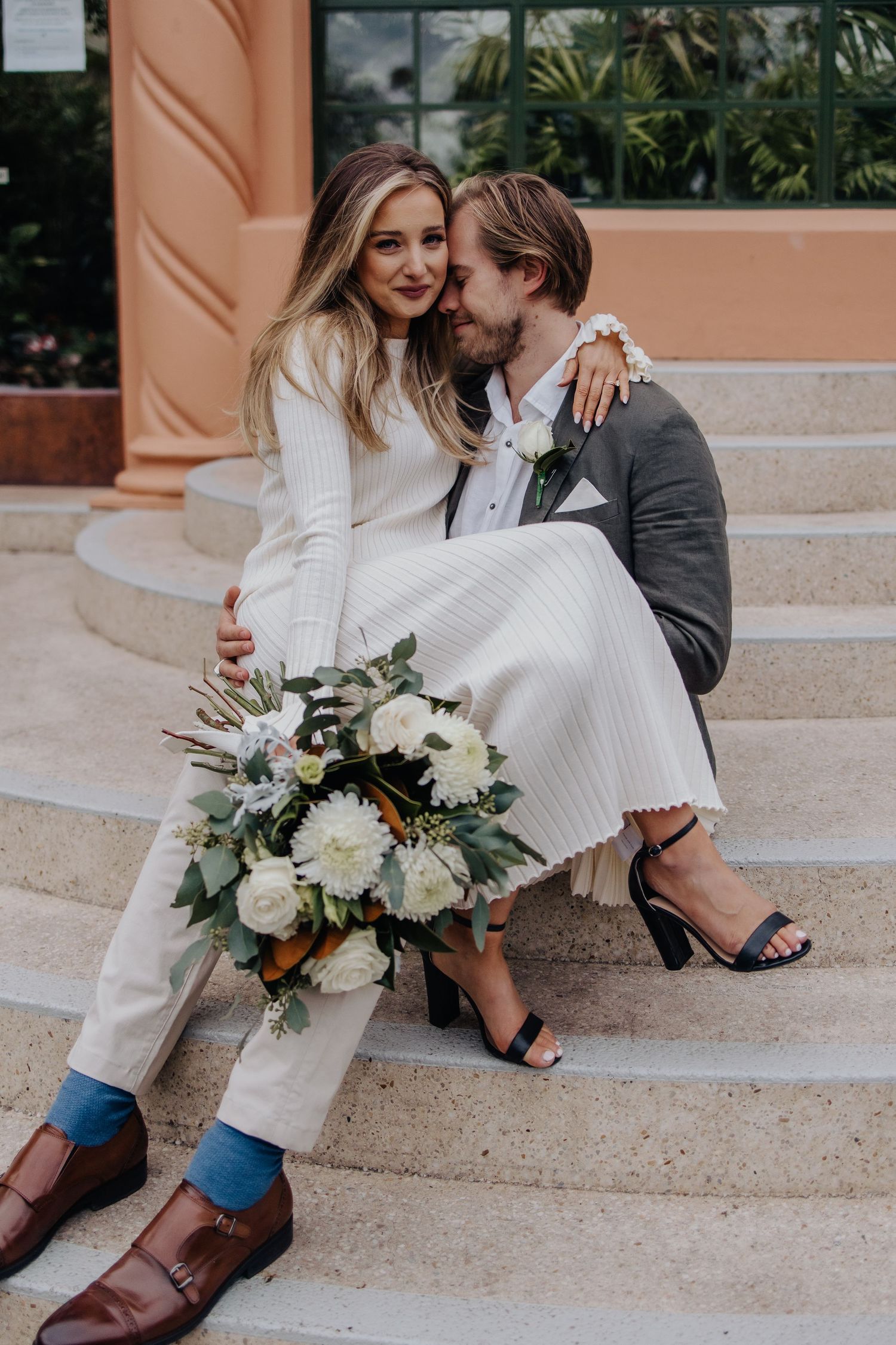 Couple in formal wear sitting on concrete steps near coral-colored building.