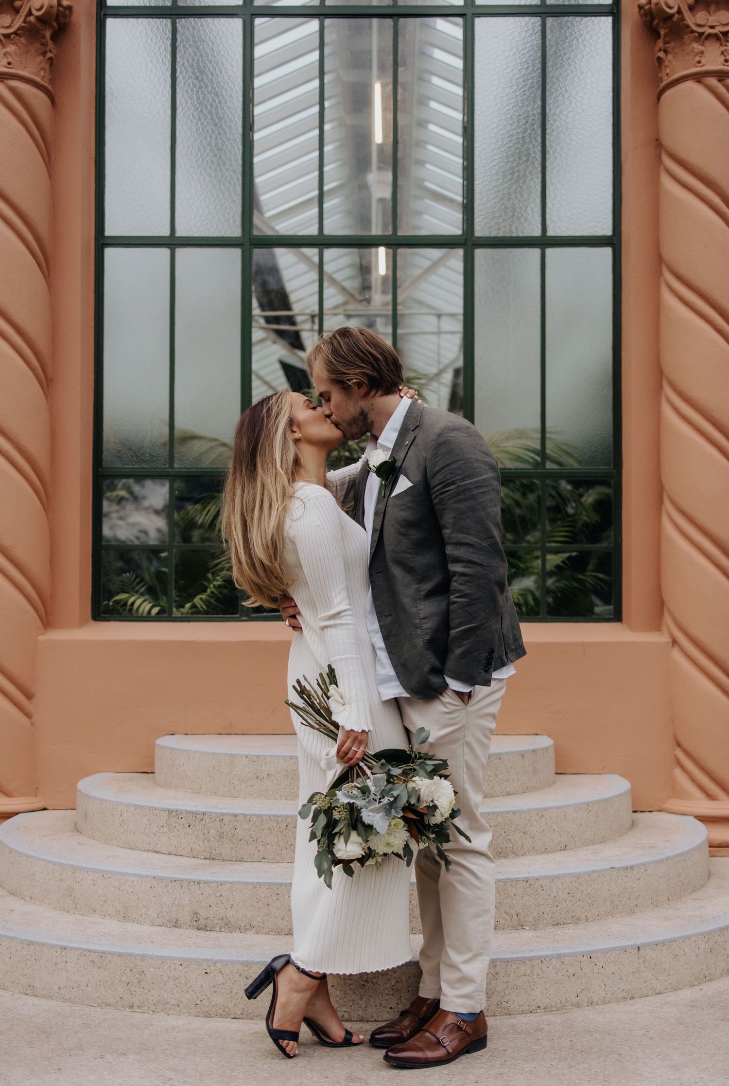A couple in formal attire embraces near stone columns and windows.