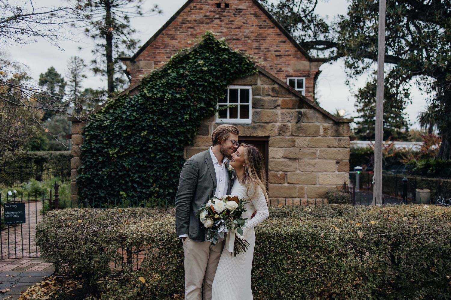 A couple poses in front of a historic brick building with ivy.