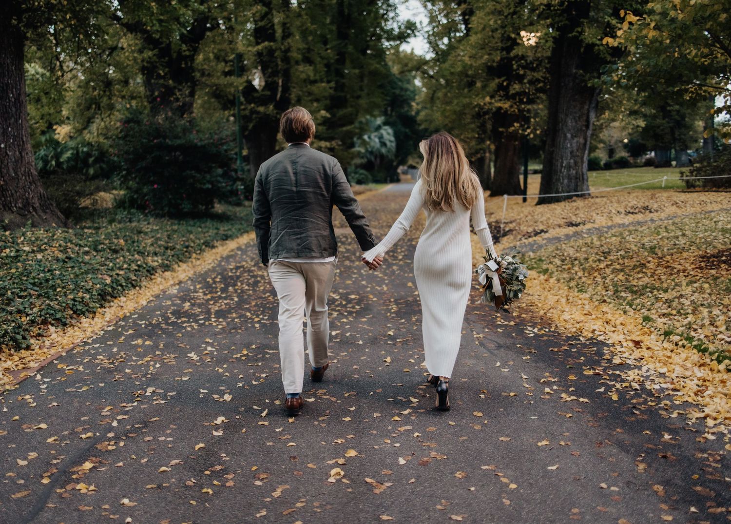 A couple walks hand in hand on a path covered in autumn leaves.