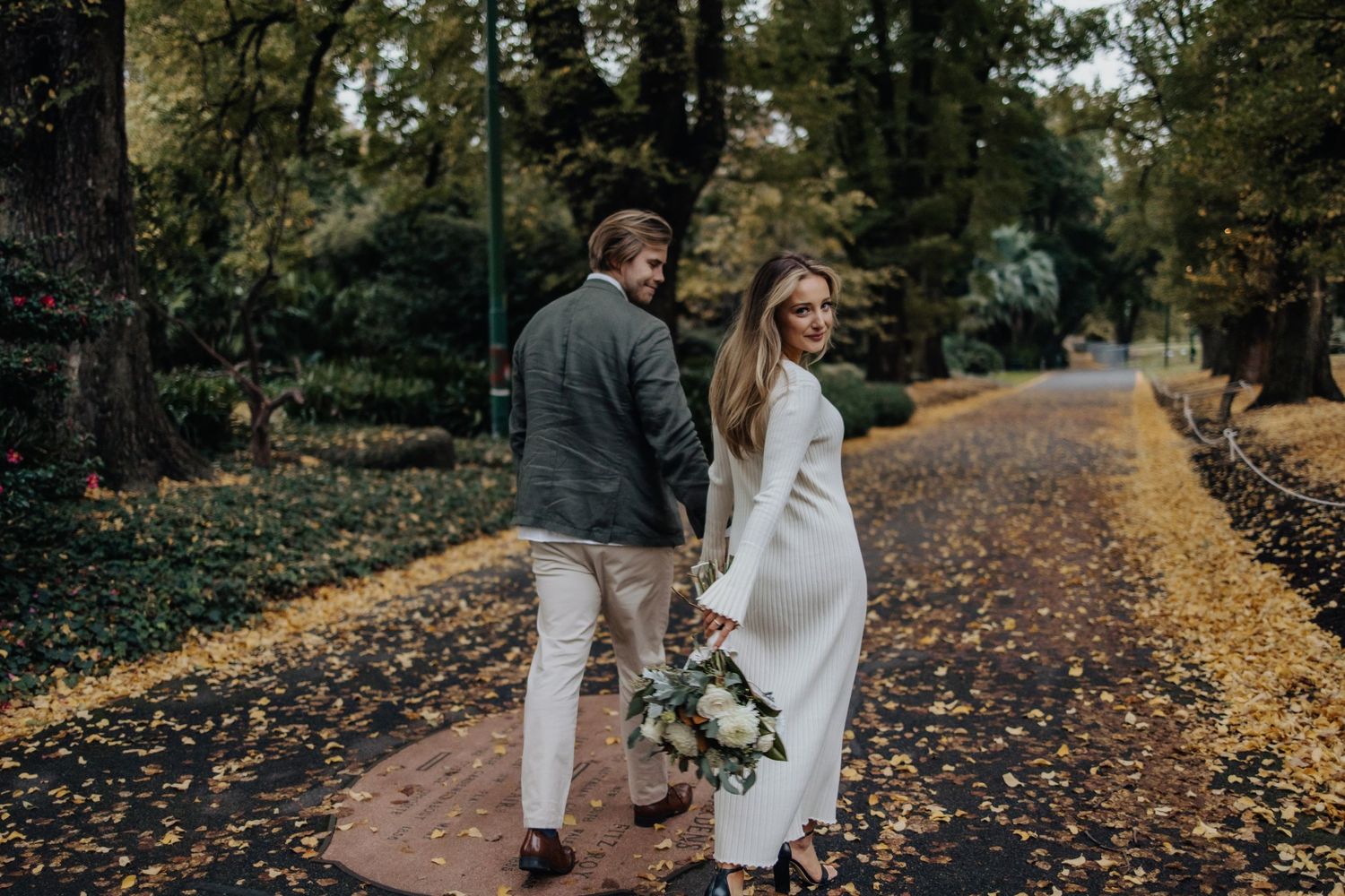 A couple strolls together on a tree-lined path with fallen leaves.