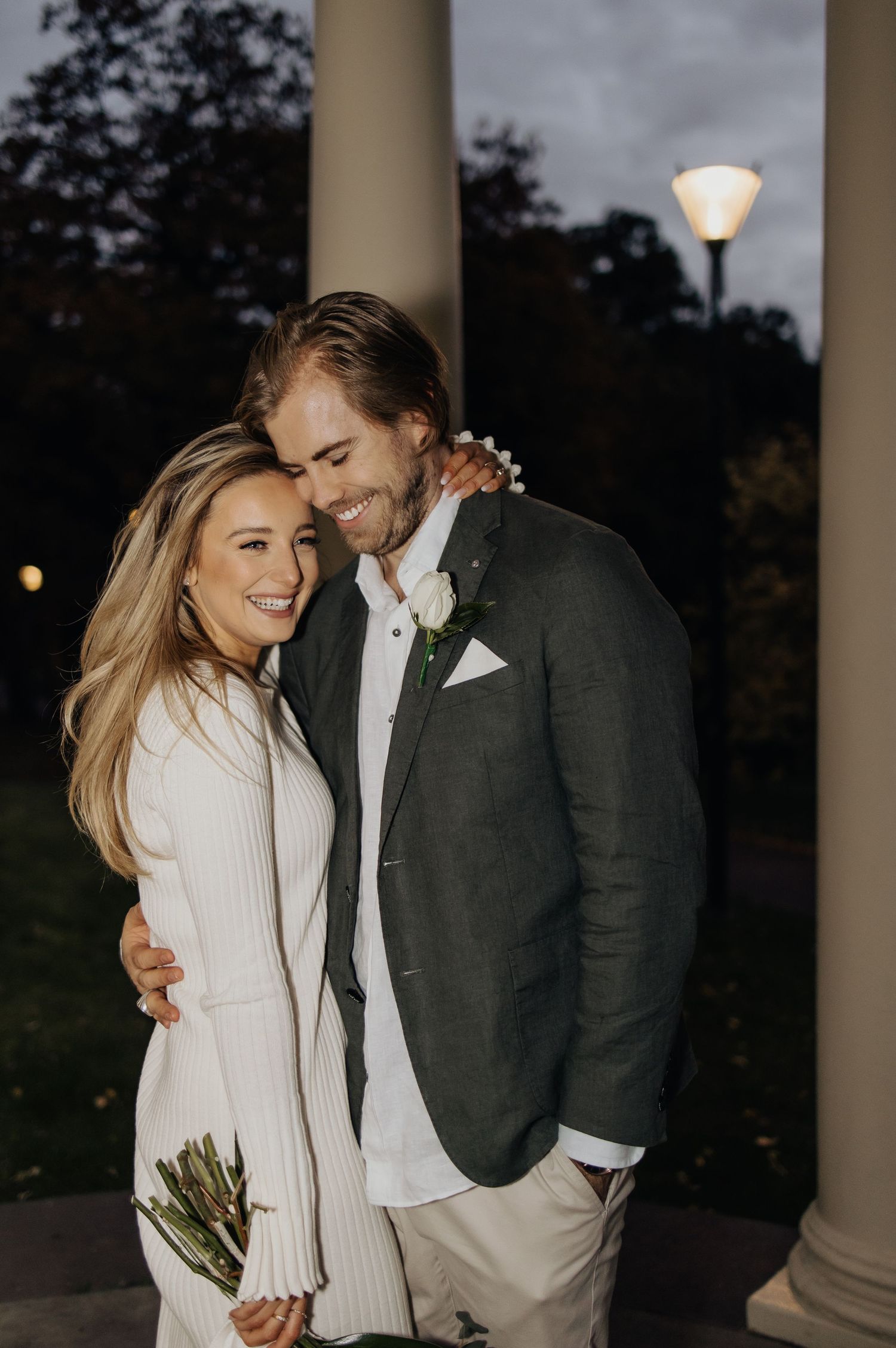 A couple in formal wear shares a tender moment under columned architecture at dusk.