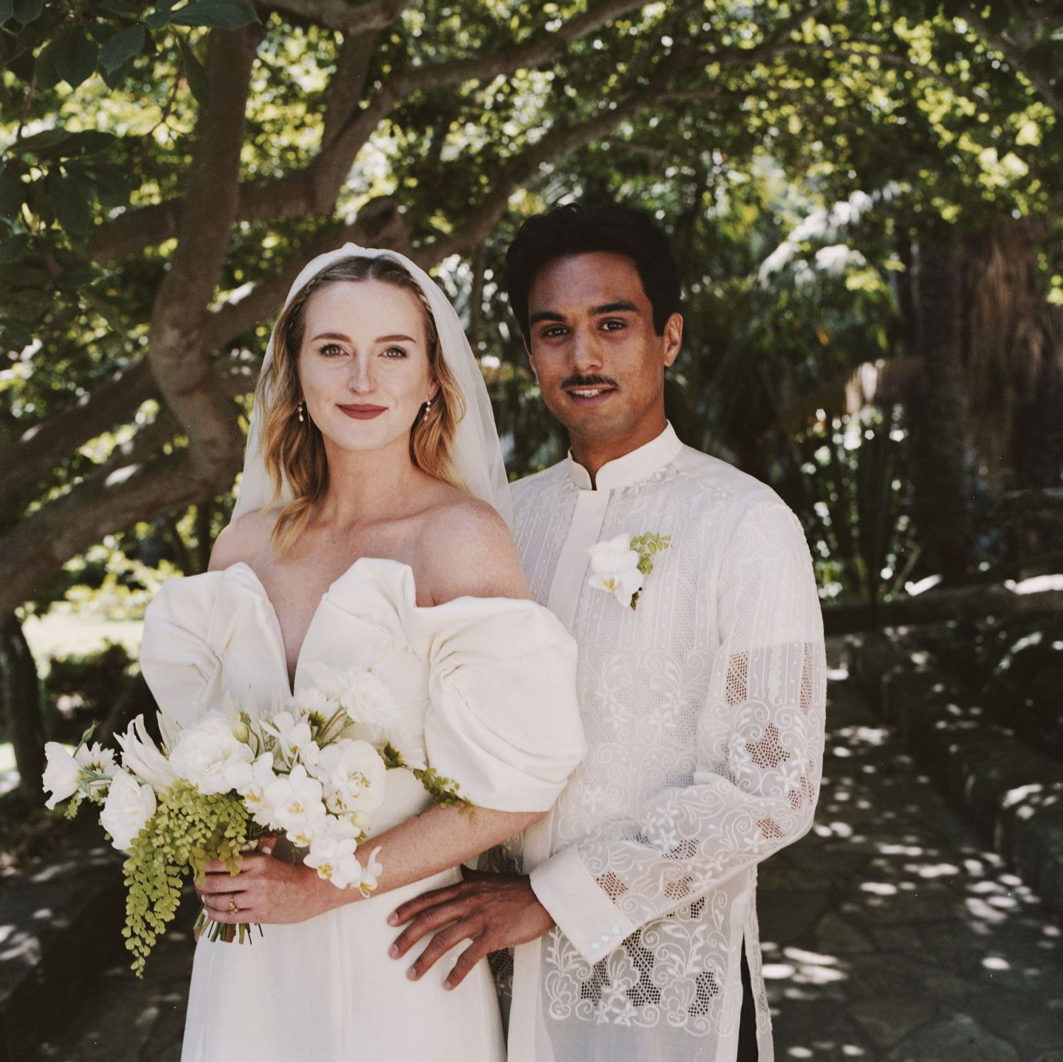 Couple in white wedding attire poses outdoors with greenery in background.
