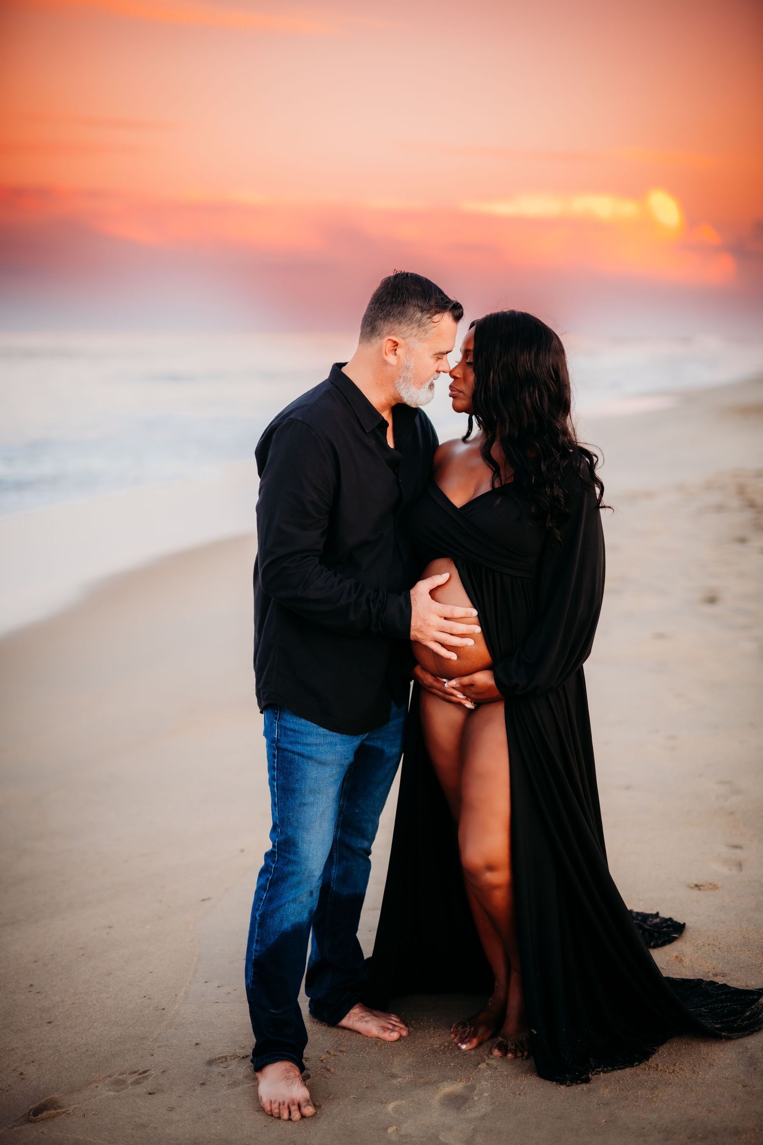 Romantic couple embraces on beach at sunset with orange sky and ocean waves in background.