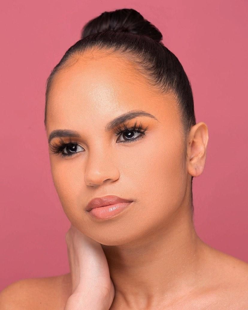 Close-up portrait of a woman with elegant makeup, orange eyeshadow, and hair in a bun on pink background.