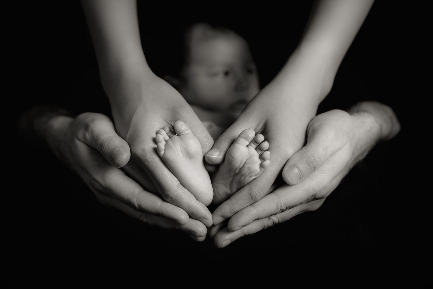 Black and white image of gentle hands cradling a tiny baby against a dark background.