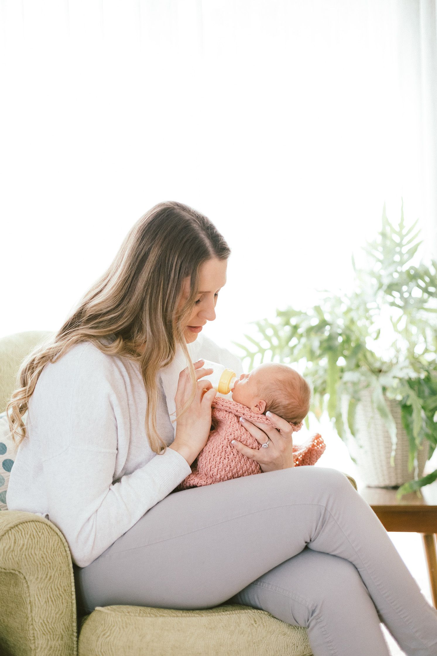 A tender moment as a caregiver bottle feeds a newborn baby wrapped in pink knit blanket while sitting in beige armchair.