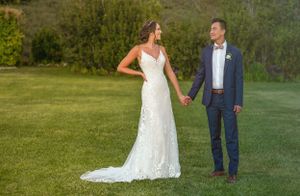 Bride in lace gown and groom in navy suit pose together on grassy lawn during wedding.