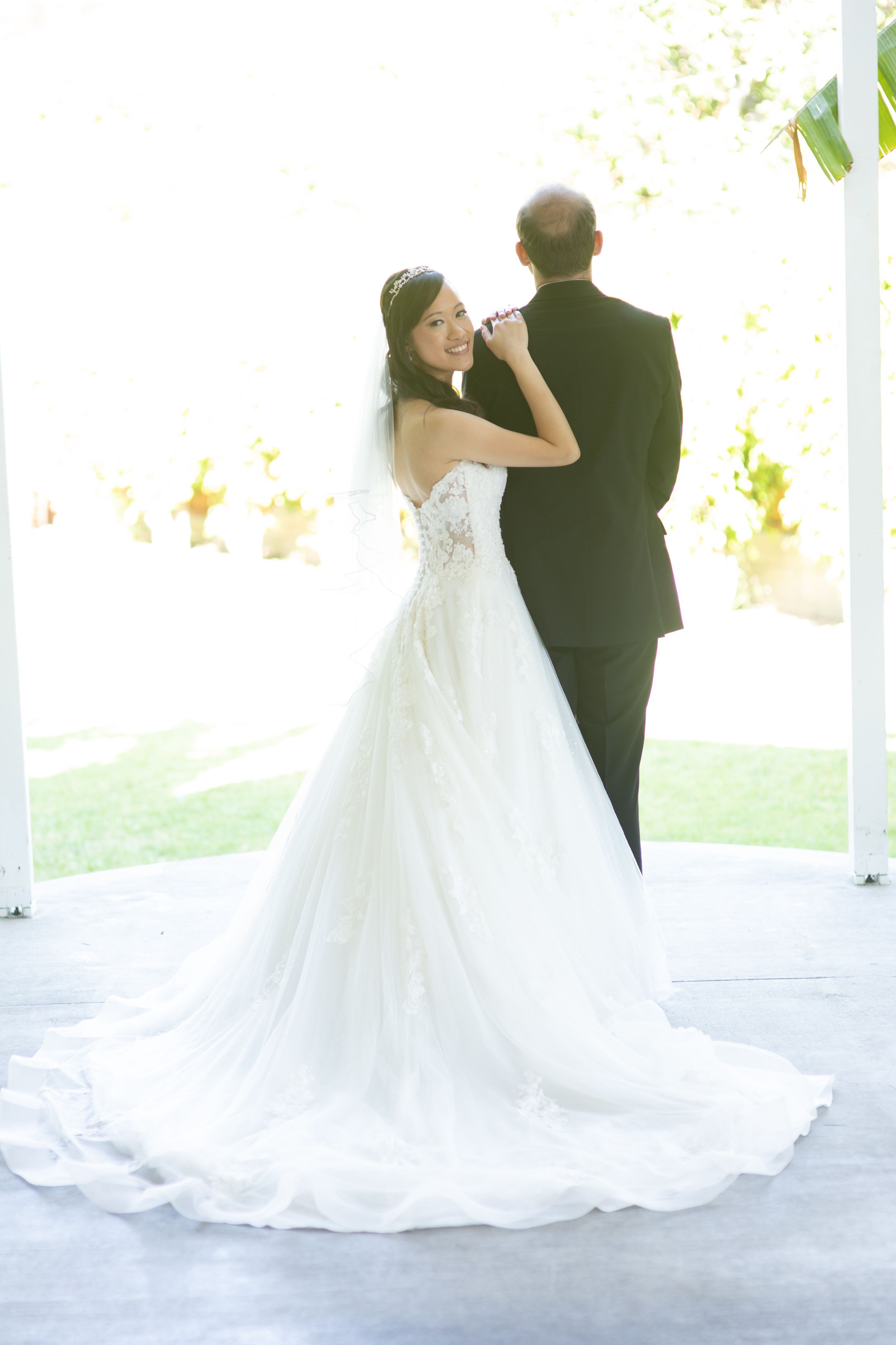 Bride in white lace gown shares intimate moment with groom in sunlit outdoor setting.