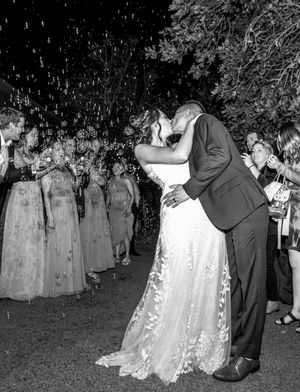 Black and white photo of wedding couple sharing romantic kiss while guests shower them with confetti.