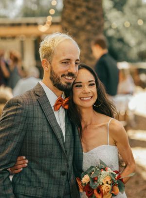 Newlyweds in grey suit and white dress smiling outdoors in warm sunlight.