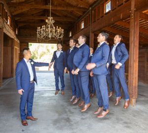 Groomsmen in navy suits and brown shoes lined up inside rustic wooden barn with chandelier.