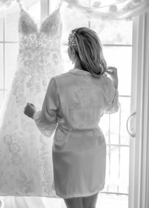 A person in a silk robe examines a hanging lace wedding dress by a window.