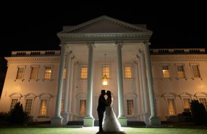A silhouetted couple stands in front of an illuminated white mansion at night.