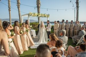 An outdoor beach wedding ceremony under a floral arch with bridesmaids in blush dresses.