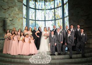 A wedding party poses for a formal group photo on stone steps in front of tall stained glass windows.