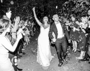 Black and white photo of newlyweds exiting reception through crowd with sparklers and raised hands.