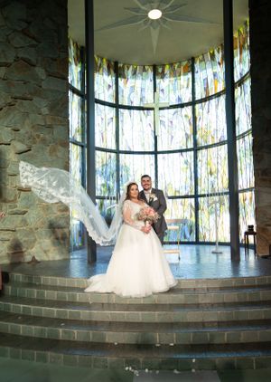 A wedding couple stands in a beautiful chapel with floor-to-ceiling stained glass windows and stone walls.